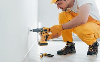 Handyman in yellow uniform works with electricity and installing new socket by using automatic screwdriver. House renovation conception photo