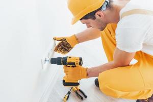 Handyman in yellow uniform works with electricity and installing new socket by using automatic screwdriver. House renovation conception photo