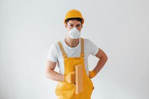 Handyman in yellow uniform and protective mask standing against white wall. House renovation conception photo