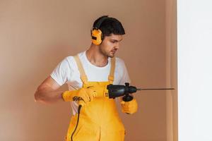 Handyman in yellow uniform works indoors by using hammer drill. House renovation conception photo