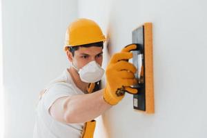 Handyman in yellow uniform and protective mask polishing wall indoors. House renovation conception photo