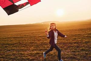 Happy little girl running with kite in hands on the beautiful field at sunrishe time photo