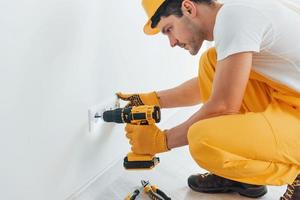 Handyman in yellow uniform works with electricity and installing new socket by using automatic screwdriver. House renovation conception photo