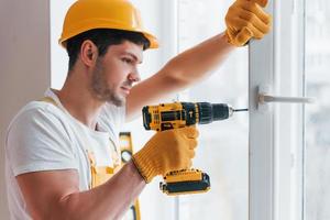 Handyman in yellow uniform installs new window by using automatic screwdriver. House renovation conception photo