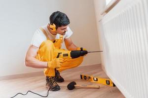 Handyman in yellow uniform works indoors by using hammer drill. House renovation conception photo