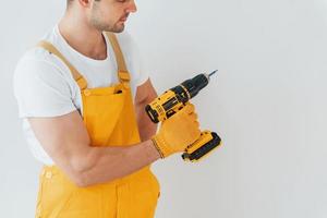 Handyman in yellow uniform standing against white wall with automatic screwdriver. House renovation conception photo