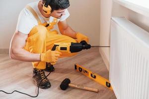 Handyman in yellow uniform works indoors by using hammer drill. House renovation conception photo