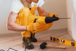 el manitas con uniforme amarillo trabaja en interiores usando un taladro percutor. concepción de renovación de la casa foto