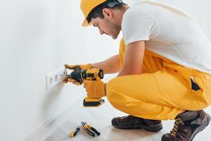 Handyman in yellow uniform works with electricity and installing new socket by using automatic screwdriver. House renovation conception photo