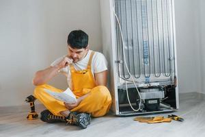 Confused handyman in yellow uniform reading manual for fridge indoors. House renovation conception photo