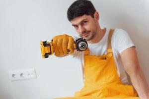 Handyman in yellow uniform standing against white wall with automatic screwdriver. House renovation conception photo