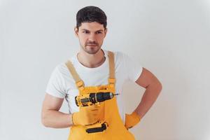 Handyman in yellow uniform standing against white wall with automatic screwdriver. House renovation conception photo