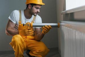 Handyman in yellow uniform works indoors with heat battery by using special tool. House renovation conception photo