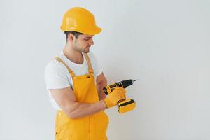 Handyman in yellow uniform standing against white wall with automatic screwdriver. House renovation conception photo