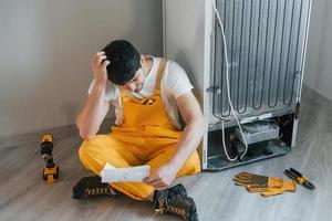 Confused handyman in yellow uniform reading manual for fridge indoors. House renovation conception photo