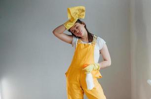 Tired housewife in yellow uniform standing with cleaning spray for windows indoors. House renovation conception photo