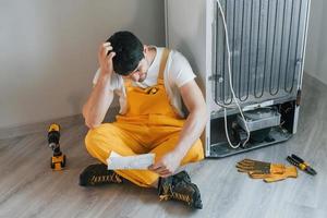 Confused handyman in yellow uniform reading manual for fridge indoors. House renovation conception photo