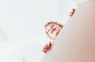 View from below of little boy with curly hair that have fun with paper box indoors photo