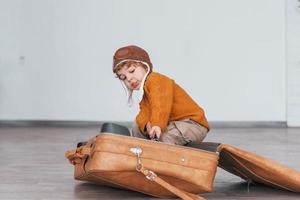 Little boys in retro pilot costume have fun and sitting in suitcase indoors at daytime photo