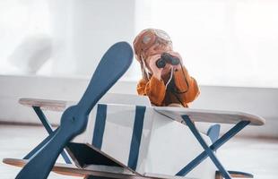 Little boy in retro pilot uniform having fun with toy plane indoors photo