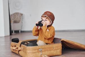 Little boys in retro pilot costume have fun and sitting in suitcase indoors at daytime photo