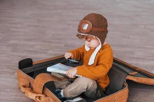 Little boys in retro pilot costume have fun and sitting in suitcase indoors at daytime photo