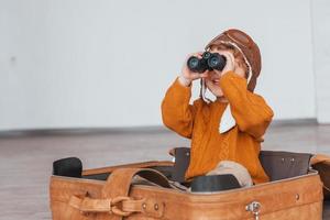 Little boys in retro pilot costume have fun and sitting in suitcase indoors at daytime photo