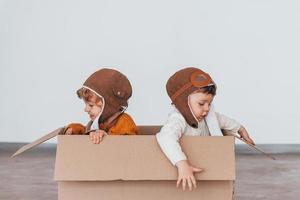 Two little boys in retro pilot costumes have fun and sitting in paper box indoors at daytime photo