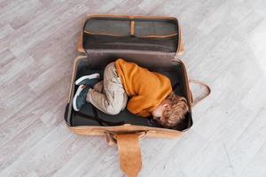 Little boys in retro pilot costume have fun and lying down in suitcase indoors at daytime photo