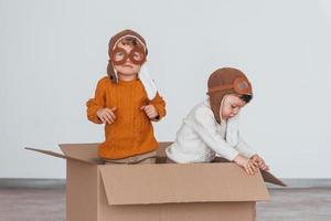 dos niños pequeños con disfraces de piloto retro se divierten y se sientan en una caja de papel en el interior durante el día foto