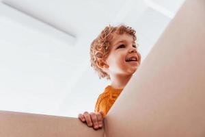 View from below of little boy with curly hair that have fun with paper box indoors photo