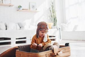 Little boys in retro pilot costume have fun and sitting in suitcase indoors at daytime photo