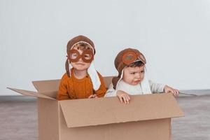 Two little boys in retro pilot costumes have fun and sitting in paper box indoors at daytime photo