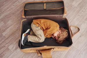 Little boys in retro pilot costume have fun and lying down in suitcase indoors at daytime photo