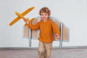 Little boy in retro pilot uniform running with toy plane indoors photo