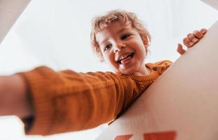 View from below of little boy with curly hair that have fun with paper box indoors photo