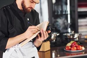 Cocinero profesional joven en uniforme de pie con bloc de notas en la cocina foto