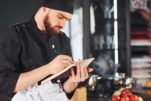 Cocinero profesional joven en uniforme de pie con bloc de notas en la cocina foto