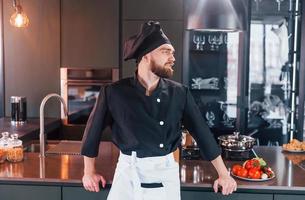 Cocinero profesional joven en uniforme de pie cerca de la mesa y posando para una cámara en la cocina foto