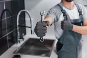 vista de cerca de un joven plomero profesional con uniforme gris sosteniendo una llave en la mano en la cocina foto