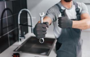 Close up view of young professional plumber in grey uniform holding wrench in hand on the kitchen photo