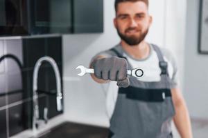 vista de cerca de un joven plomero profesional con uniforme gris sosteniendo una llave en la mano en la cocina foto