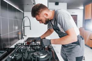 Young professional plumber in grey uniform working on the kitchen photo