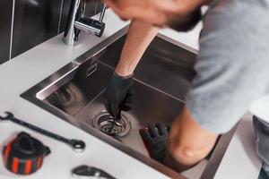Young professional plumber in grey uniform working on the kitchen photo