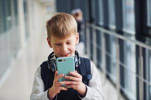 niño pequeño con teléfono y auriculares de pie frente a los niños de la escuela en uniforme que juntos en el pasillo. concepción de la educación foto