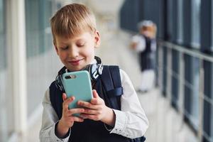 Little boywith phone and headphones standing if front of school kids in uniform that together in corridor. Conception of education photo