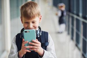 Little boywith phone and headphones standing if front of school kids in uniform that together in corridor. Conception of education photo