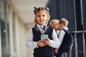 Little girl with phone standing if front of school kids in uniform that together in corridor. Conception of education photo