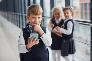 Little boywith phone and headphones standing if front of school kids in uniform that together in corridor. Conception of education photo
