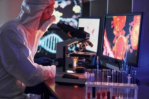 monitores con información sobre la mesa. científico con uniforme protector blanco trabaja con coronavirus y tubos de sangre en laboratorio foto